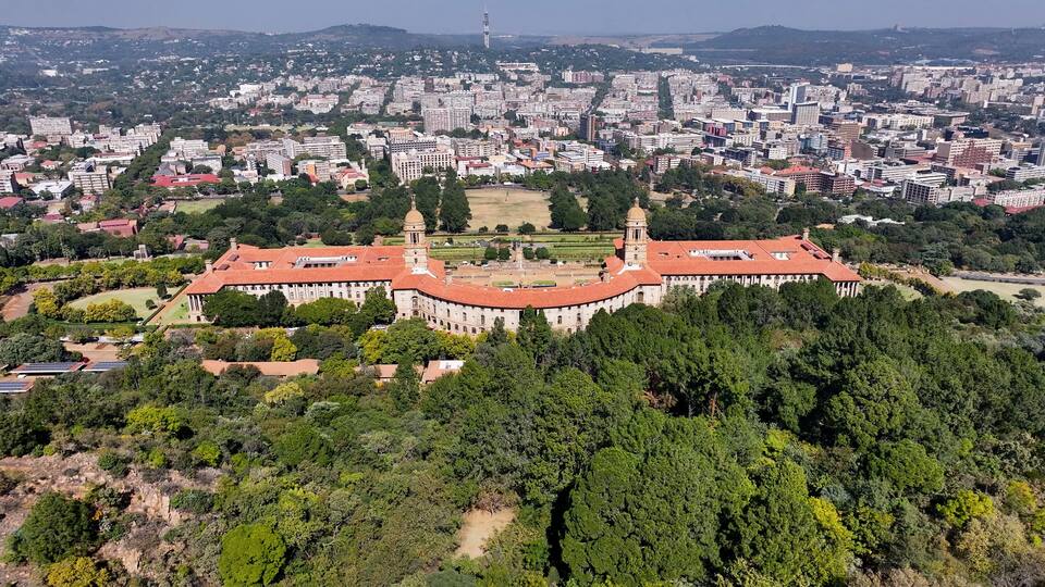A large historical building with a red roof, surrounded by dense greenery and a sprawling cityscape in the background.