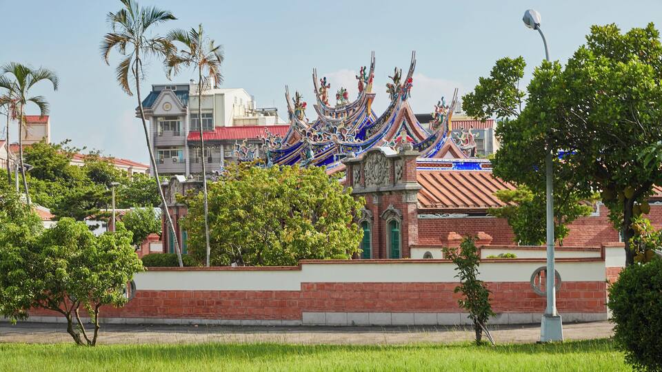 Ein traditionelles Gebäude mit aufwendigen blau-roten Dächern, umgeben von Grünflächen und modernen Bauten.