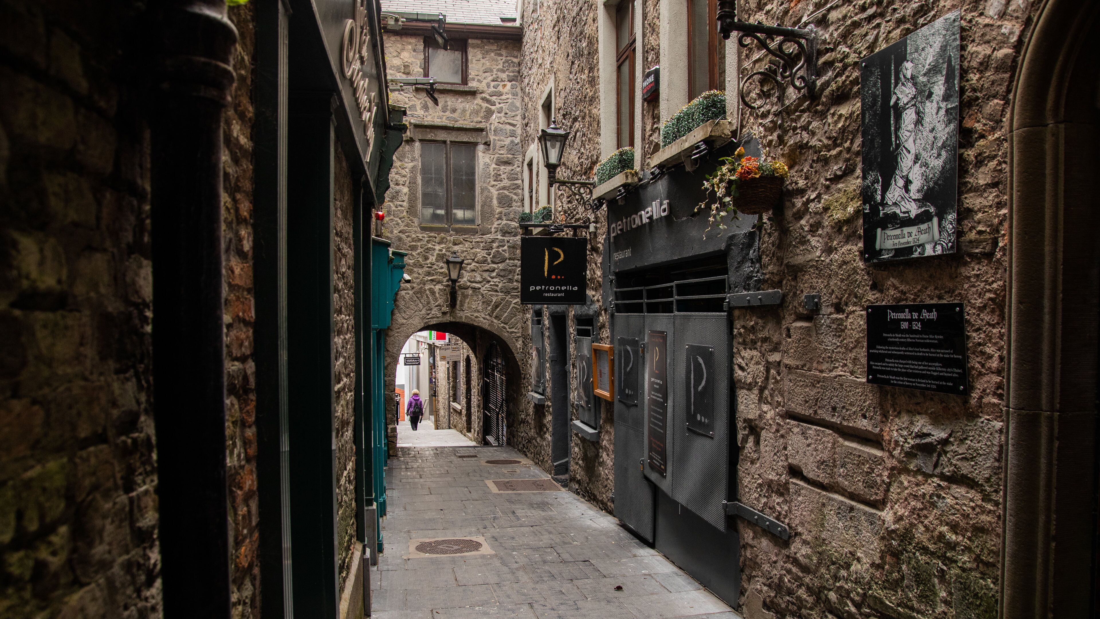 Un estrecho callejón con edificios de piedra, un letrero de restaurante y una placa conmemorativa histórica.