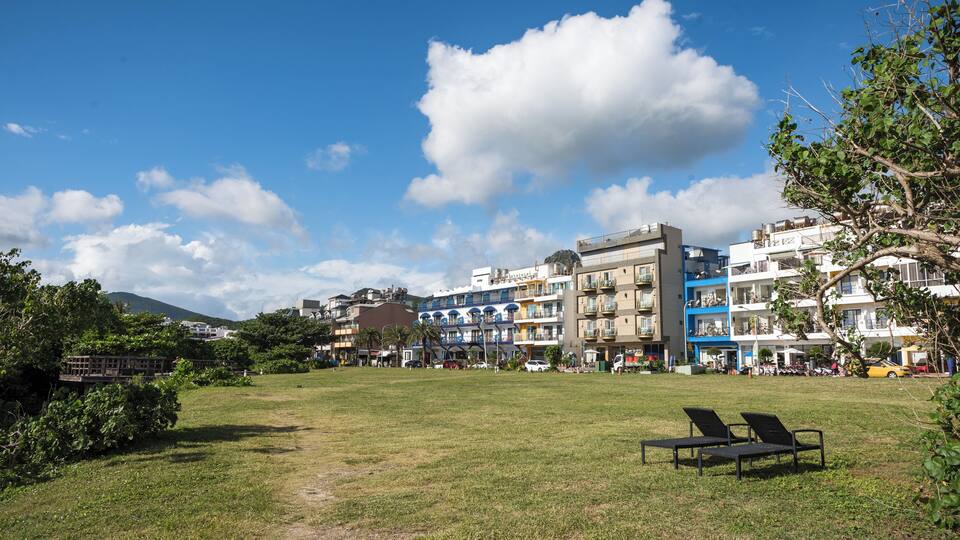 Un espacio con césped, dos sillas de descanso, un edificio de varios pisos con balcones y un cielo azul despejado con nubes.