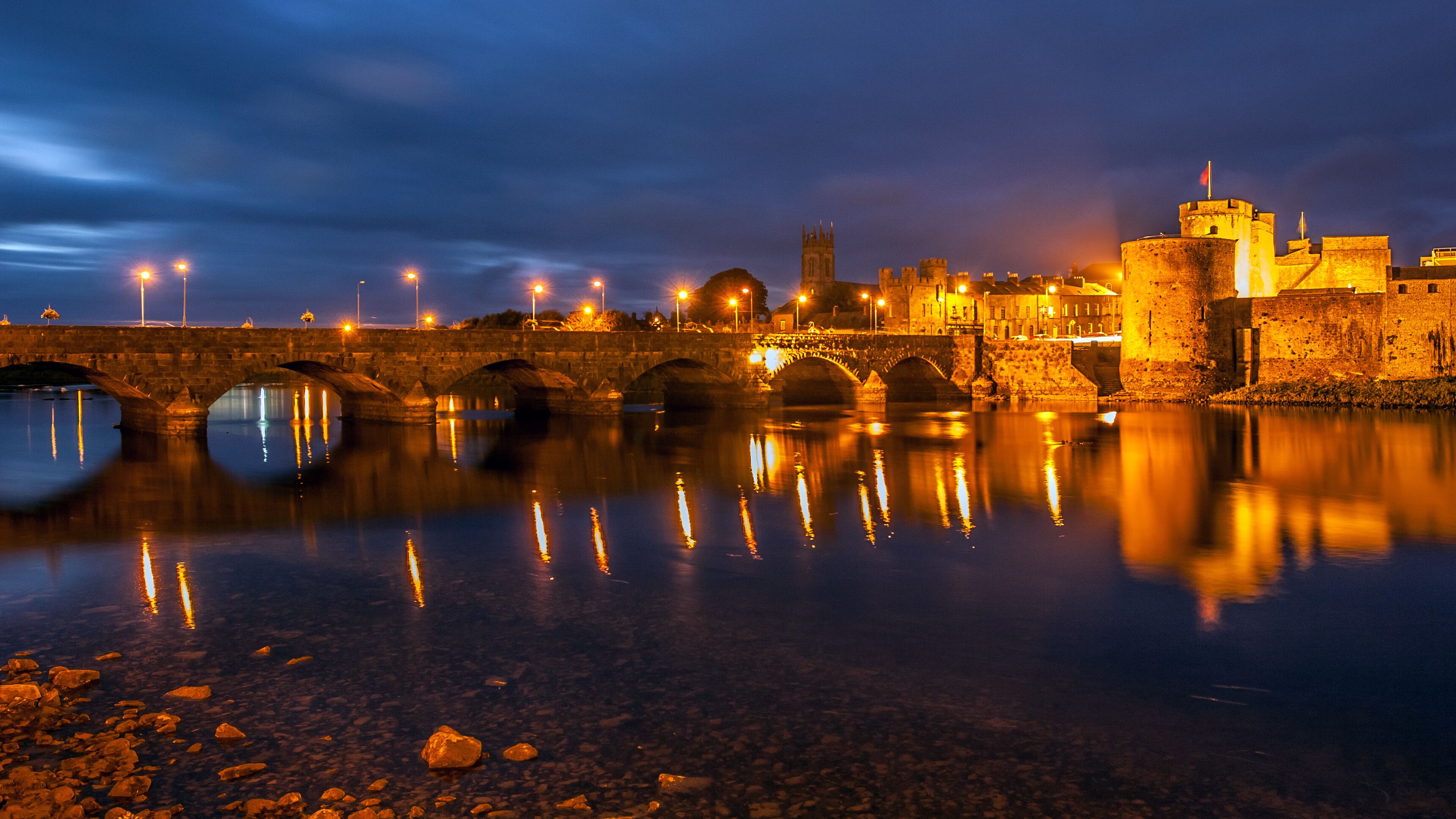 A stone bridge over a calm river reflecting city lights, with a castle and church in the background.