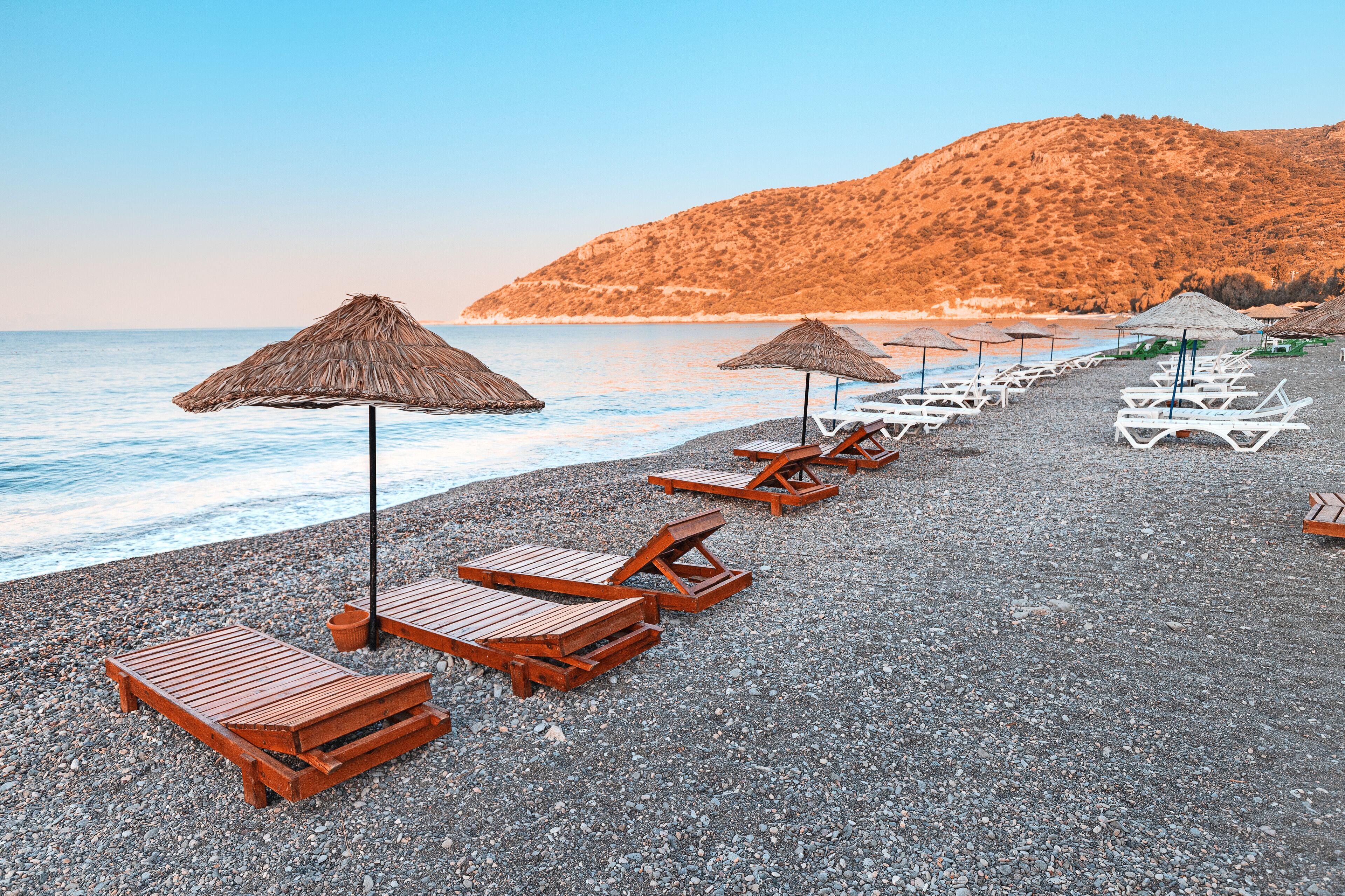 A pebble beach with wooden lounge chairs and thatched umbrellas.