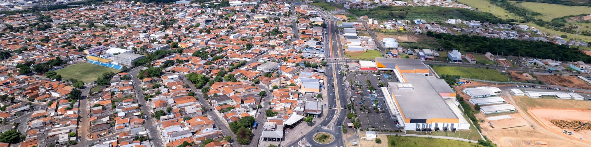 An aerial view of a densely populated urban area with a mix of residential and commercial buildings, a prominent highway, and surrounding green fields.