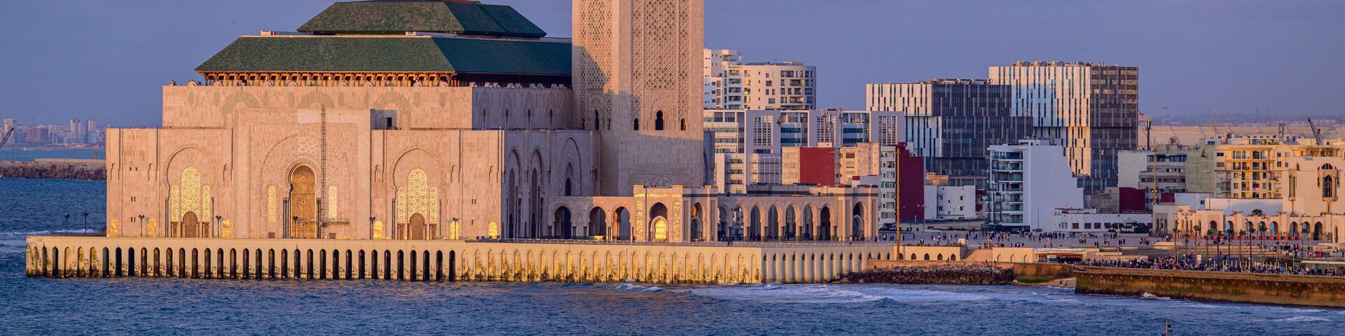 A historic mosque with a tall minaret and a large dome, situated by the sea with modern buildings in the background.