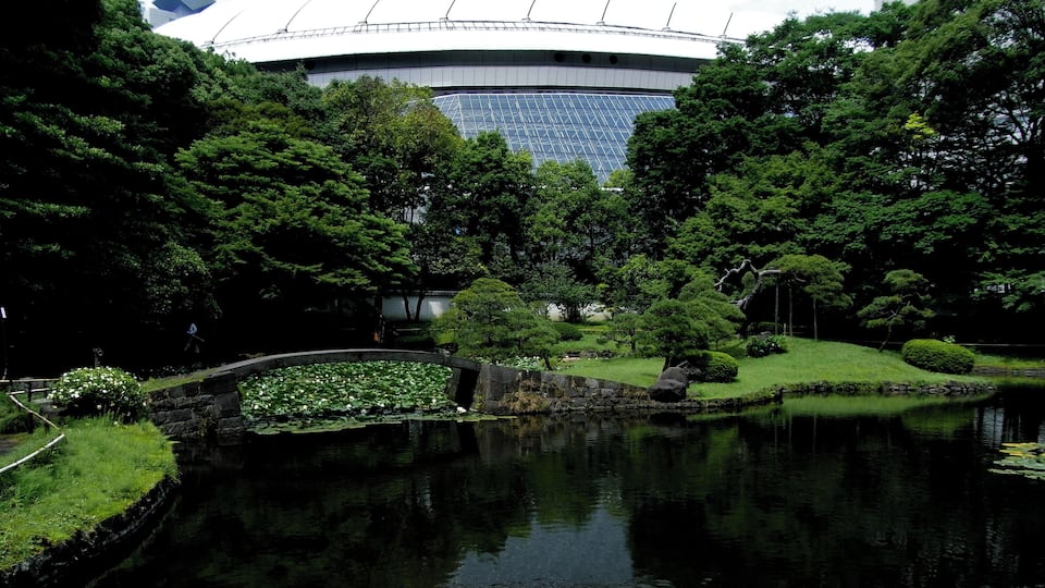 Un parc paisible avec un étang, une végétation luxuriante et, au loin, un stade moderne.