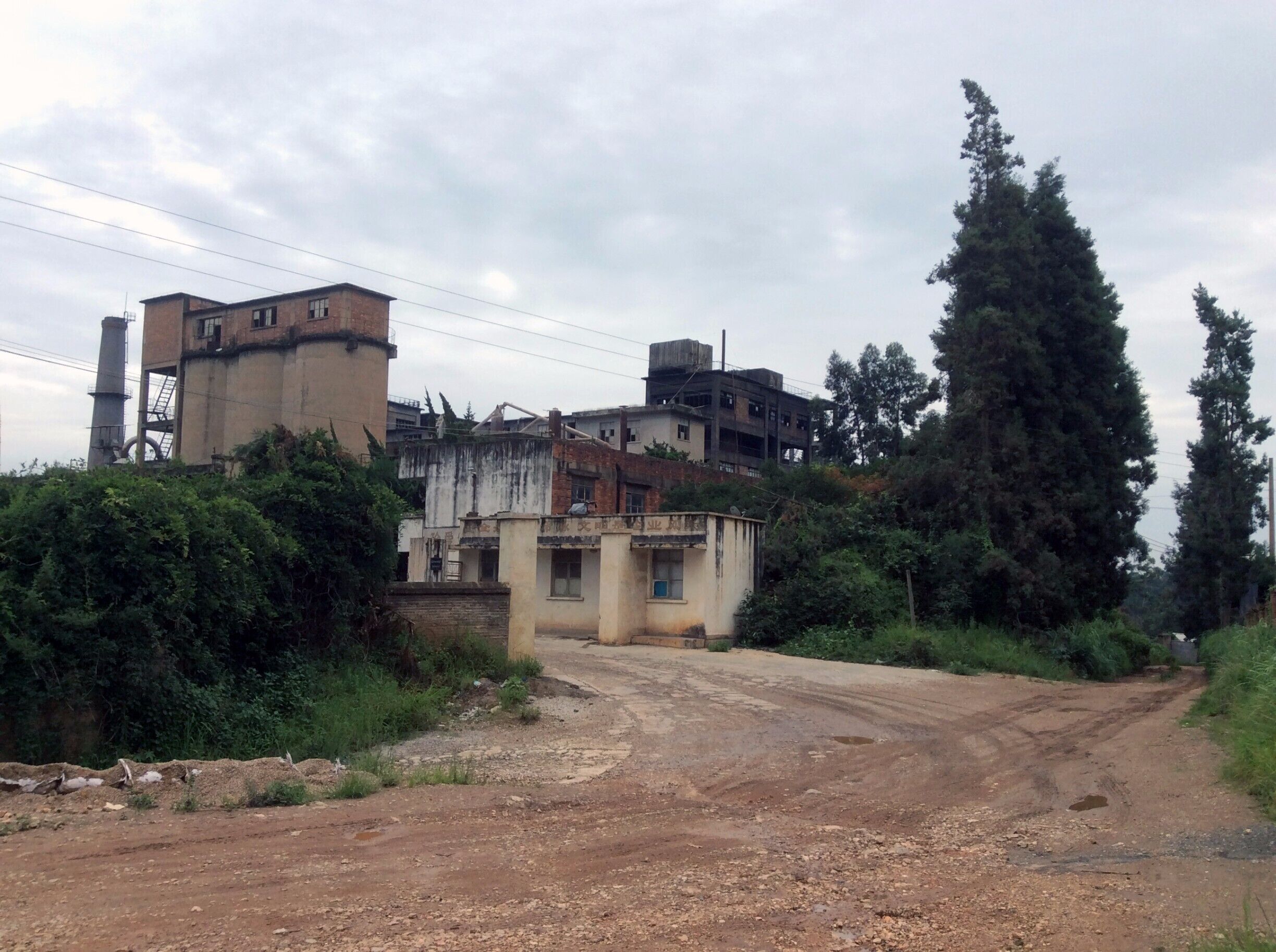 A dirt road leading to a cluster of buildings with a tall chimney and a large tree.