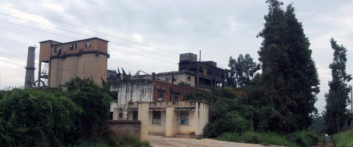A dirt road leading to a cluster of buildings with a tall chimney and a large tree.