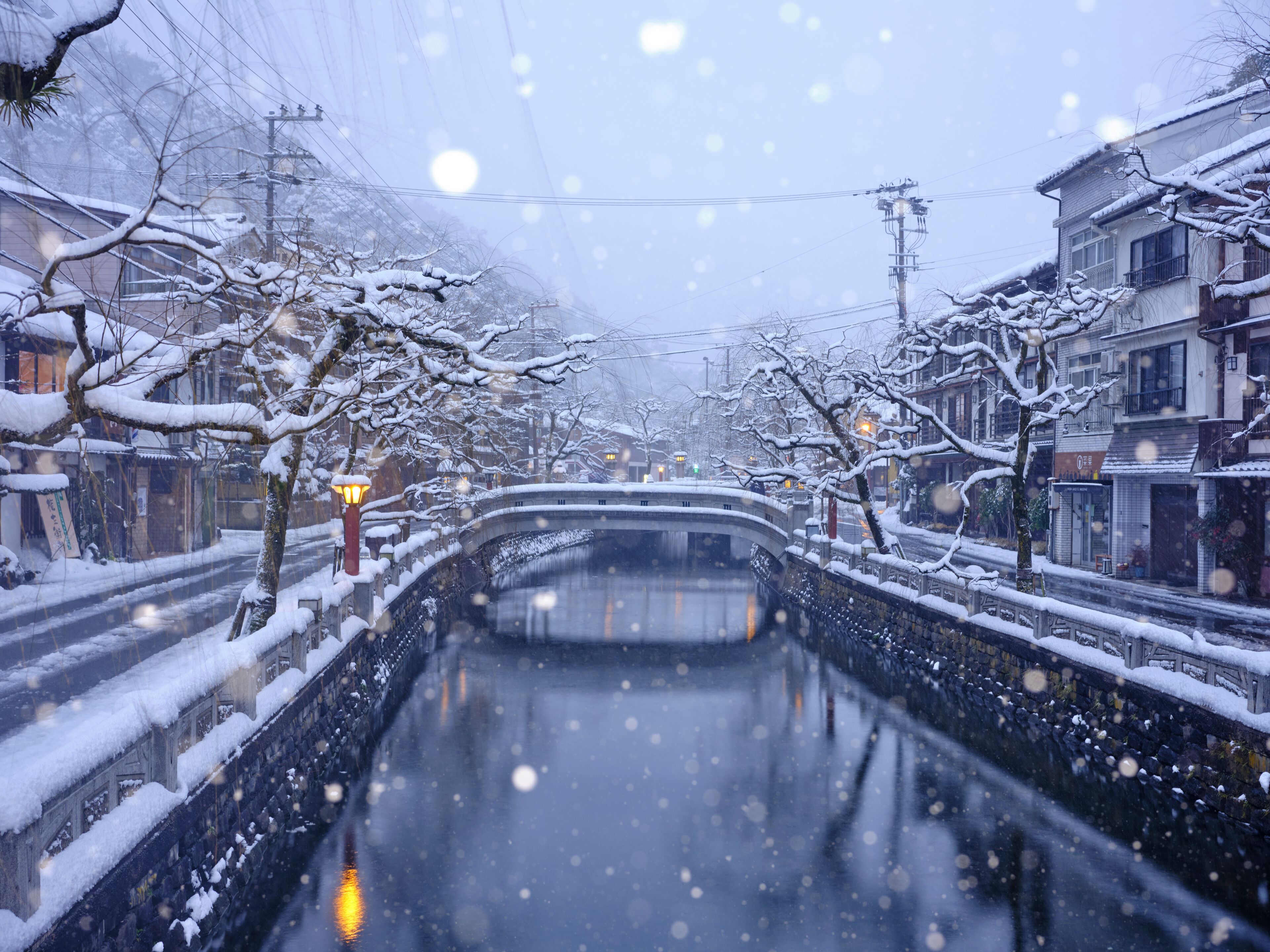A snow-covered canal with buildings on either side, a bridge in the distance, and falling snowflakes.