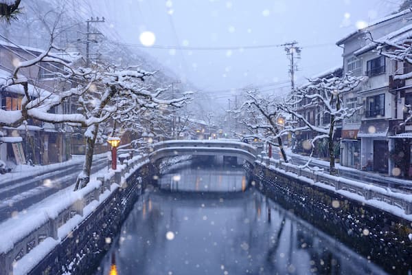 Un canal enneigé, bordé de bâtiments de chaque côté, un pont à l’horizon et des flocons de neige qui tombent.