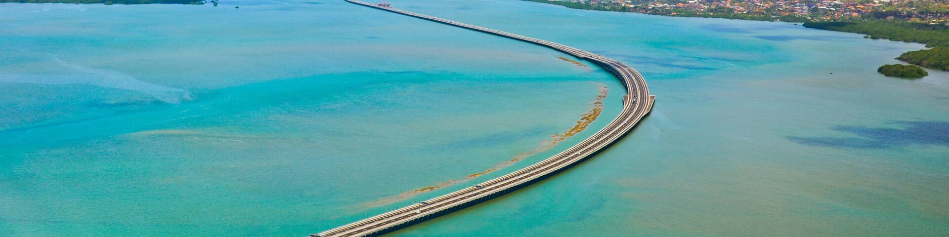 An aerial view of a long, curved bridge extending into a turquoise sea, surrounded by a coastal cityscape.