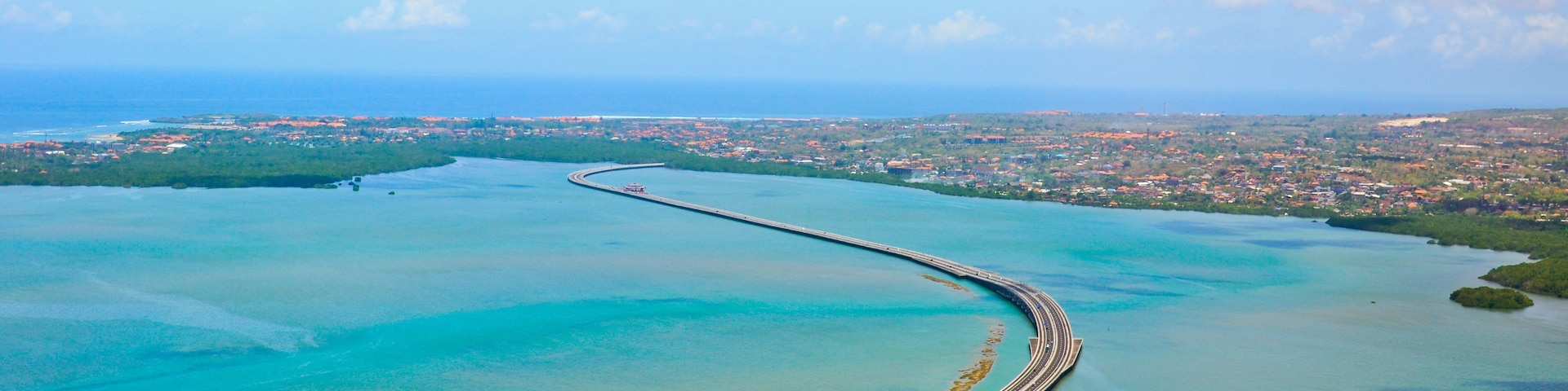 An aerial view of a long, curved bridge extending into a turquoise sea, surrounded by a coastal cityscape.