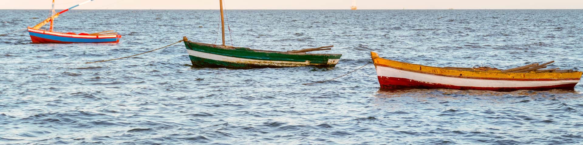 Three small boats anchored on calm water with a distant ship visible on the horizon.