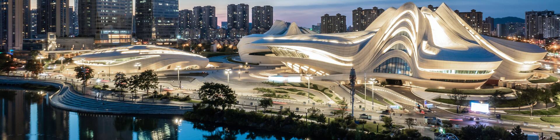 A modern architectural complex with a unique curved design, surrounded by high-rise buildings and a body of water reflecting the structures.
