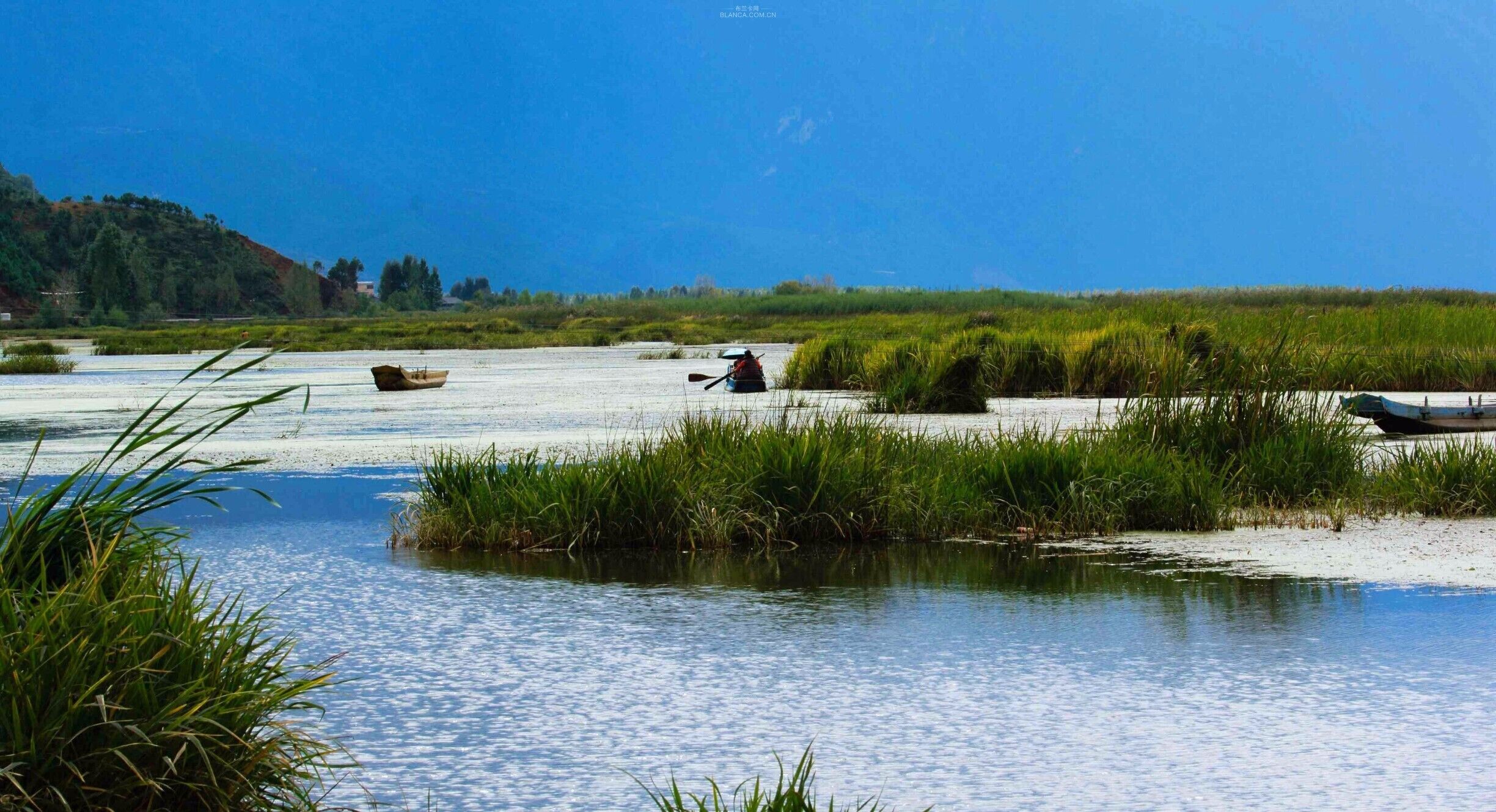 A calm river with boats, surrounded by grassy banks and a distant hill.