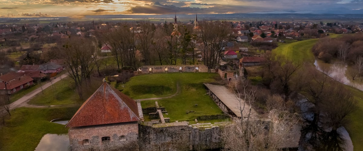 Um castelo histórico cercado por um fosso, com uma igreja ao fundo e uma cidade do outro lado do rio.