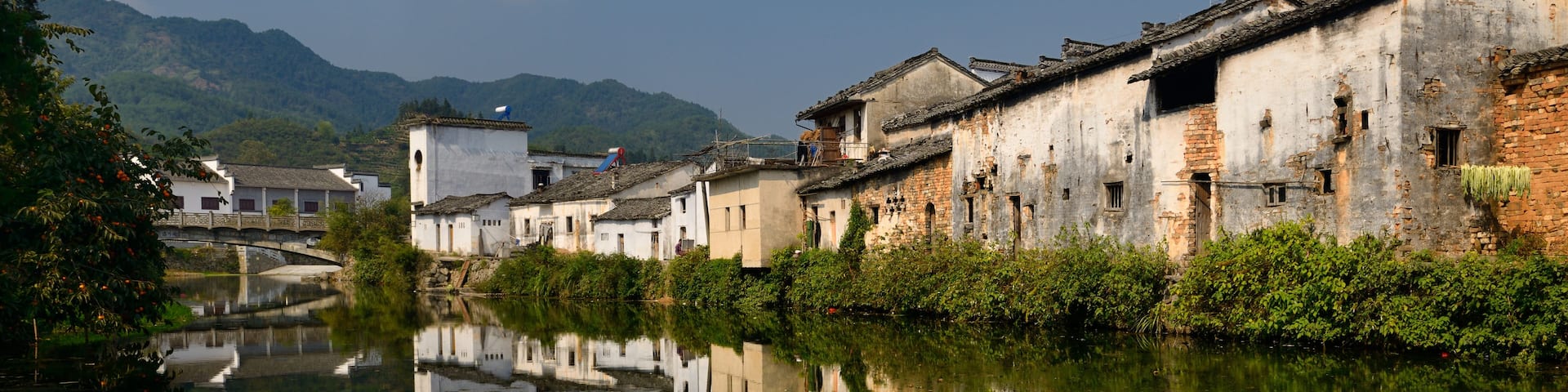A serene riverside with traditional buildings reflecting in the water, surrounded by lush greenery and distant hills.