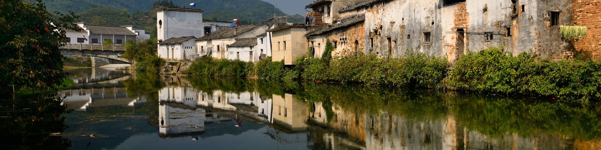 Um rio calmo com edifícios tradicionais refletidos na água, cercado por vegetação exuberante e colinas ao longe.