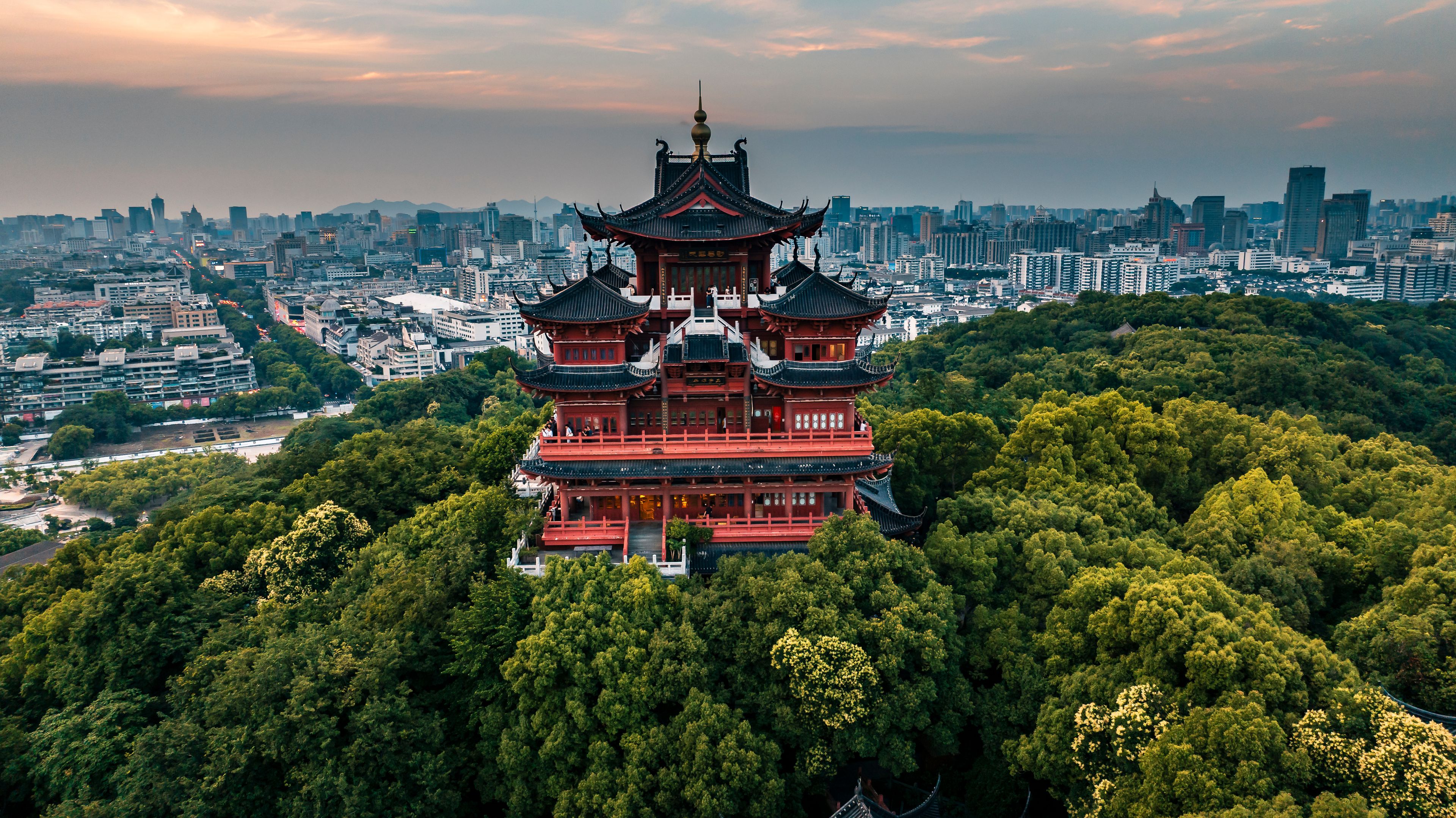 Aerial sunset scenery of Chenghuang Pavilion in Hangzhou, red ancient Chinese pavilion surrounded by green woods, West Lake and urban cityscape distant view at dusk
