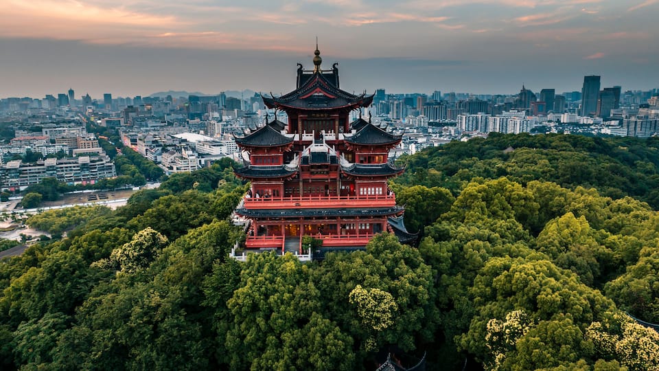 Aerial sunset scenery of Chenghuang Pavilion in Hangzhou, red ancient Chinese pavilion surrounded by green woods, West Lake and urban cityscape distant view at dusk