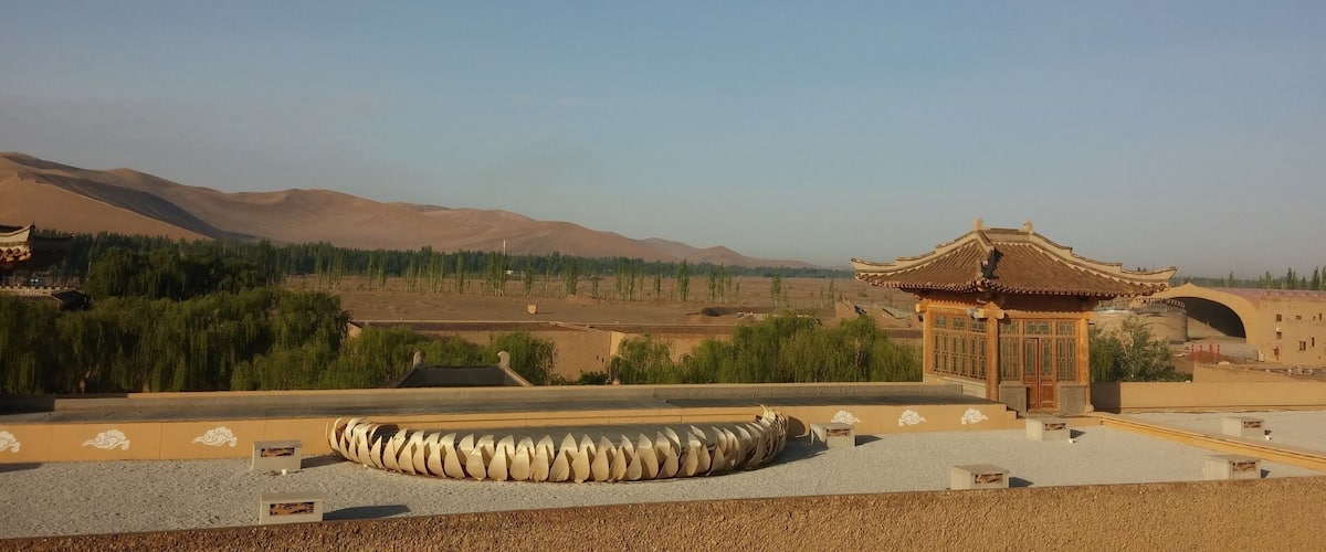 A traditional pavilion with a tiled roof, surrounded by a gravel area with a decorative stone spiral. In the background, there are mountains and a forested area.