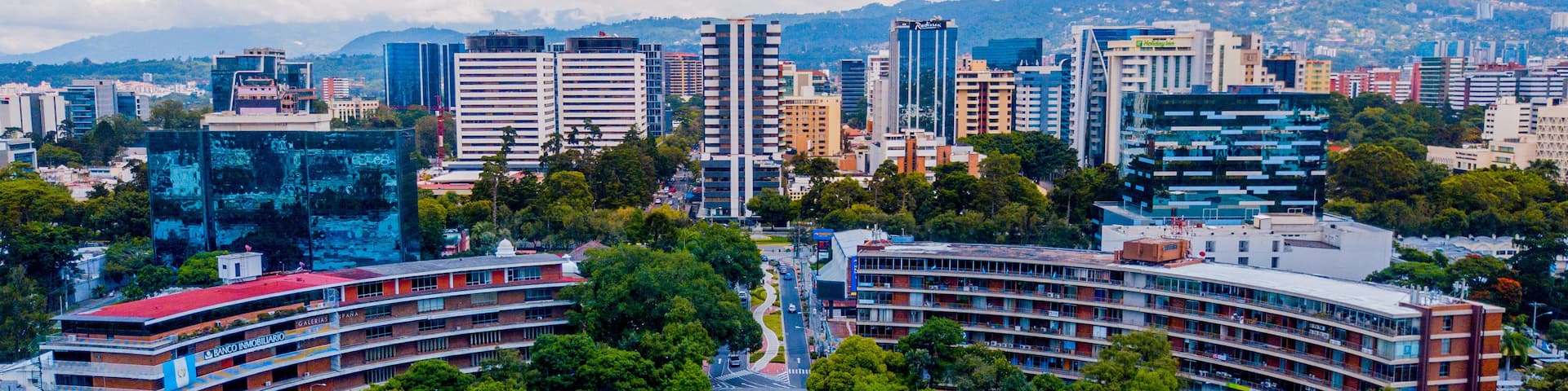 A cityscape with a central roundabout, surrounded by modern buildings and lush greenery.
