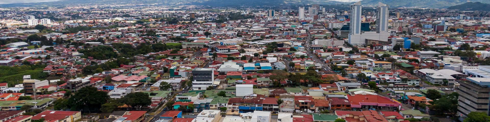 Paisagem urbana com edifícios residenciais e comerciais, um edifício azul de destaque e montanhas ao fundo.