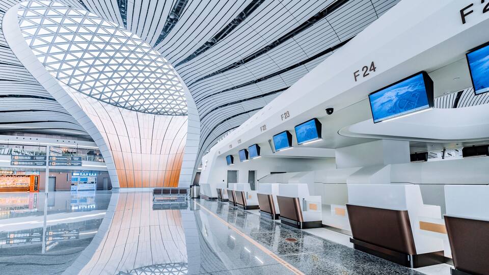 A modern airport terminal with a unique geometric ceiling design, multiple flight information screens, and a clean, reflective floor.