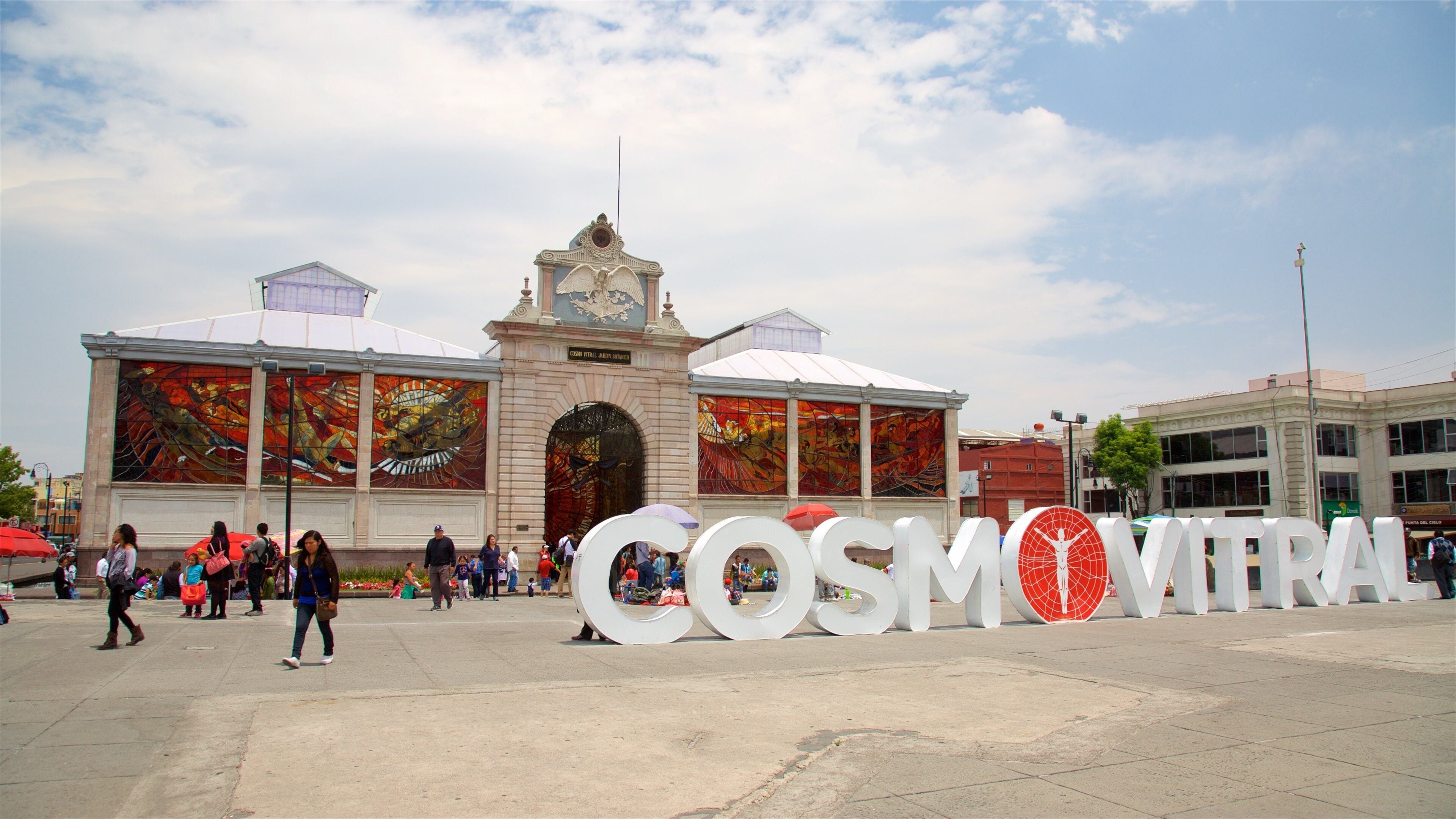 A large outdoor space with a prominent building featuring a clock tower and large windows. In front of the building, there are large white letters spelling out "COSMOTRIAL".