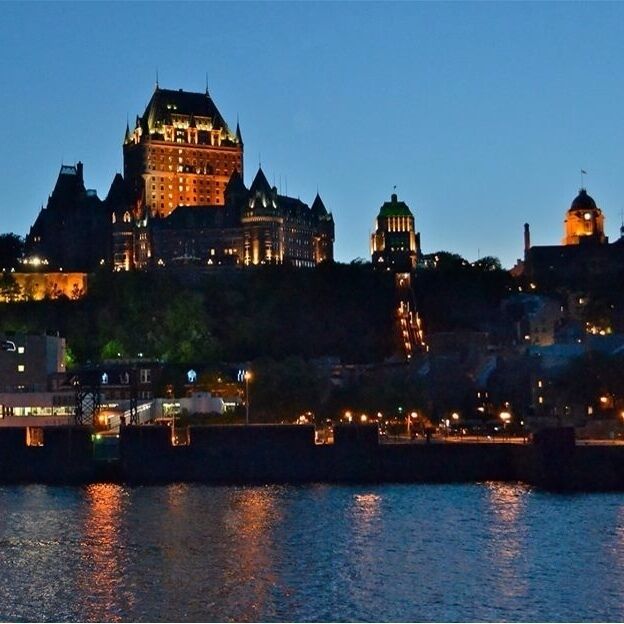 A historic castle with multiple towers and a large central building, surrounded by water and other buildings.