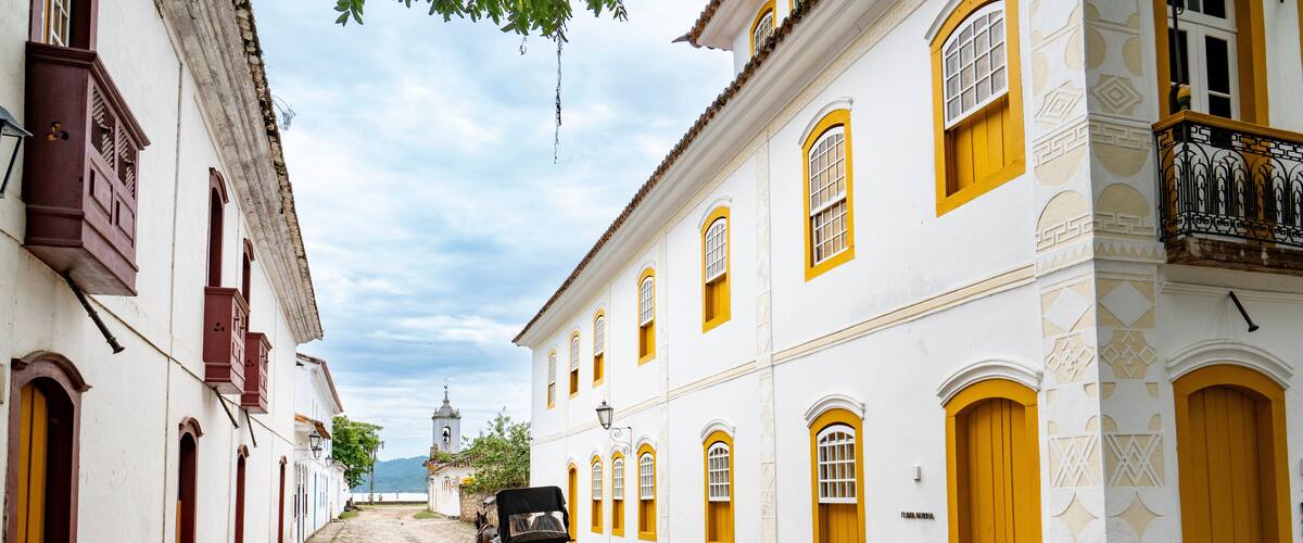 An empty historic cobblestone street featuring white colonial buildings with bright yellow windows and a horse-drawn carriage in Paraty, Brazil.