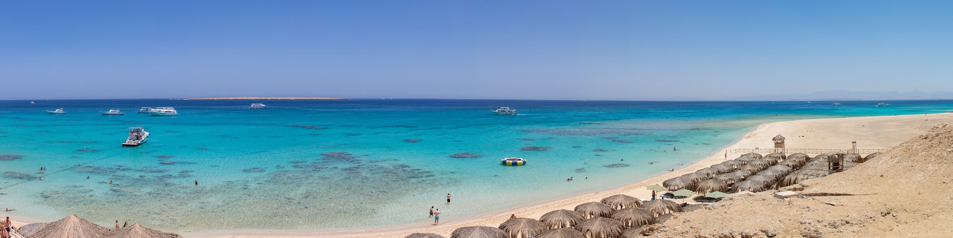 A beach with clear turquoise waters, boats, and thatched umbrellas.