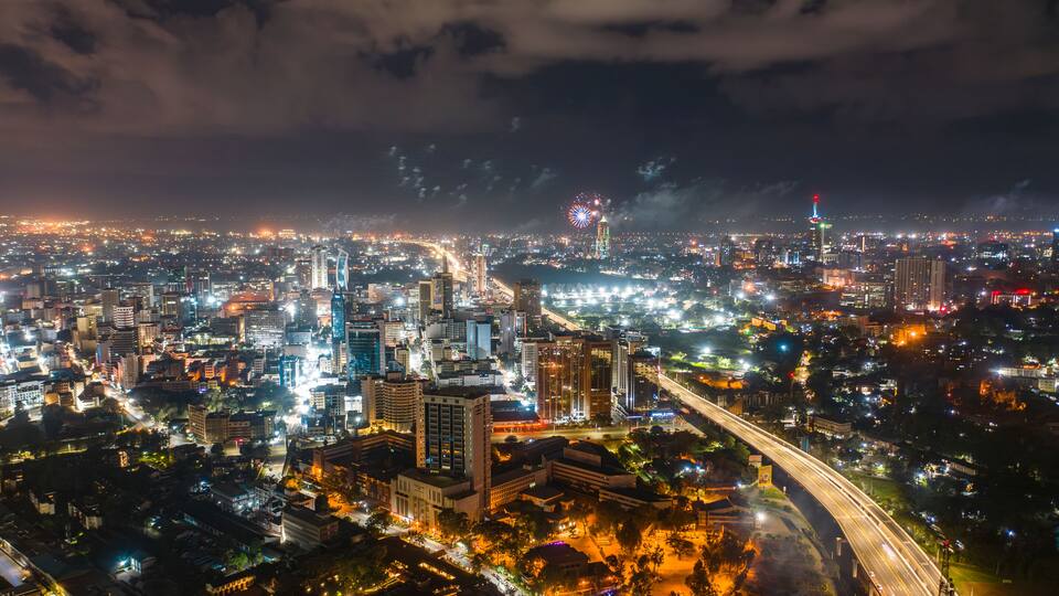 A nighttime cityscape with illuminated buildings, a prominent bridge, and fireworks in the sky.