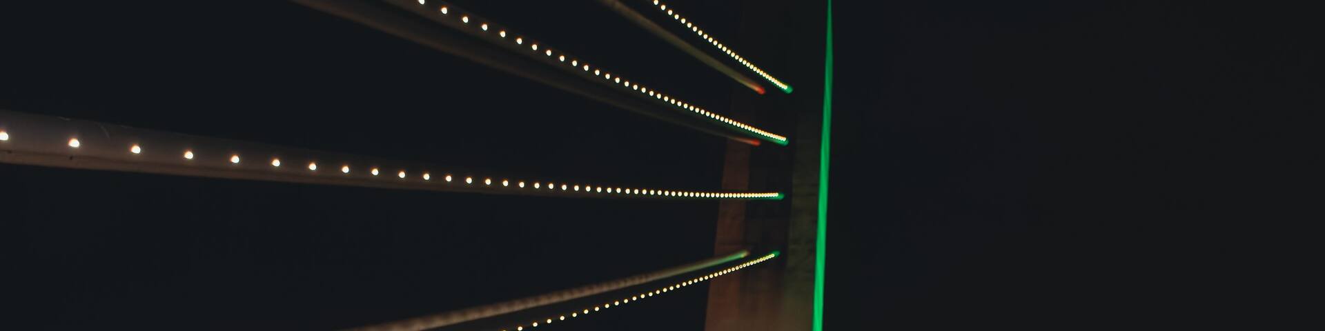 A brightly lit bridge with a unique triangular design, featuring illuminated strings of lights and a green vertical light strip.