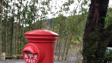 Un buzón rojo con caracteres japoneses y la palabra "POST", situado cerca de un árbol y una carretera.