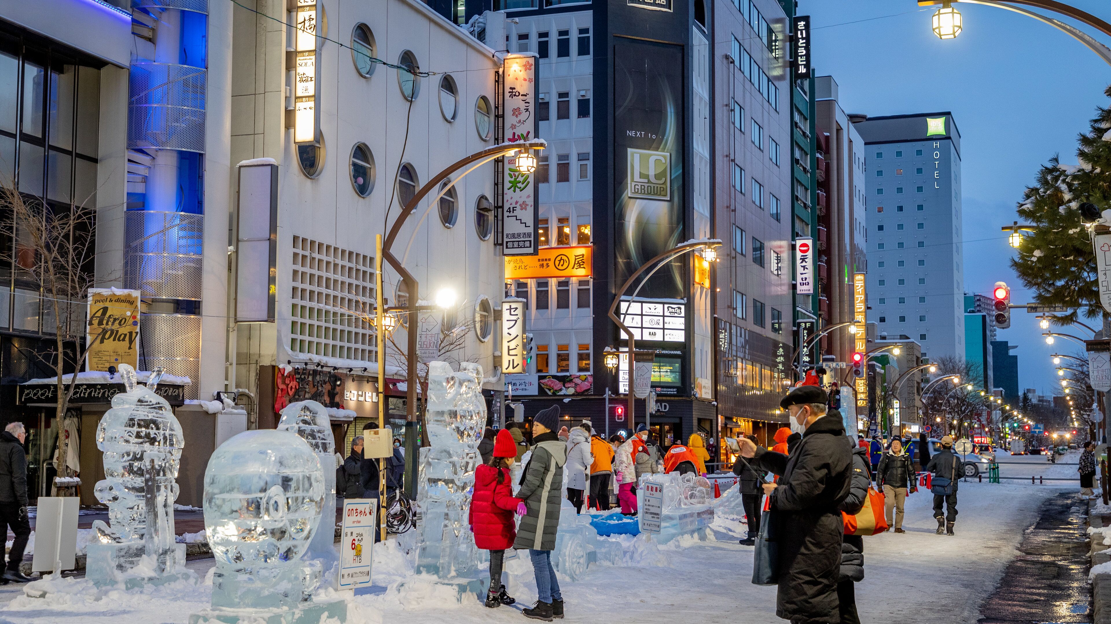A snowy urban street with ice sculptures, shops, and illuminated signs.