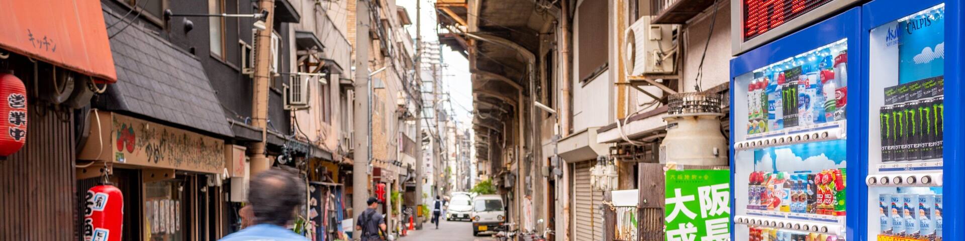 A narrow urban street with bicycles parked on the side, a vending machine, and traditional Japanese signage.