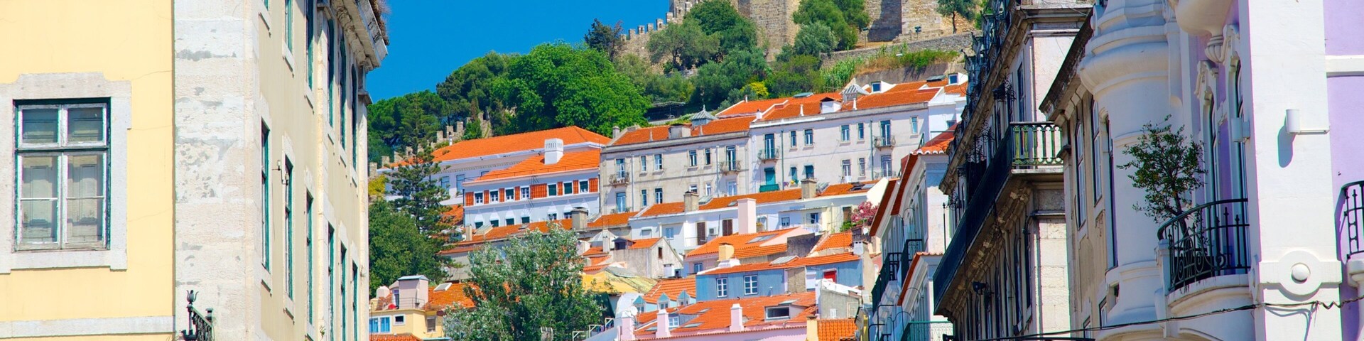 Uma cidade histórica com edifícios coloridos, um castelo imponente numa colina e um céu azul límpido.