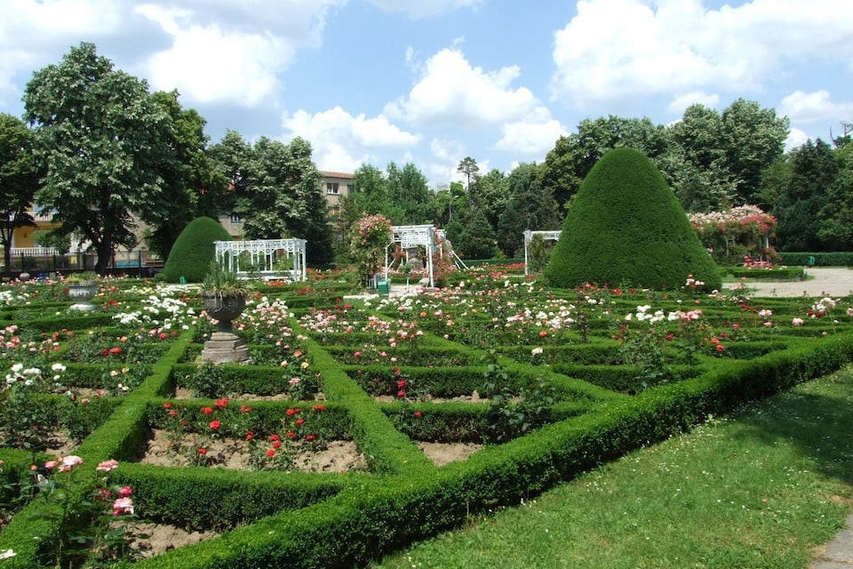 A formal garden with neatly trimmed hedges, a variety of flowering plants, and a central white gazebo.