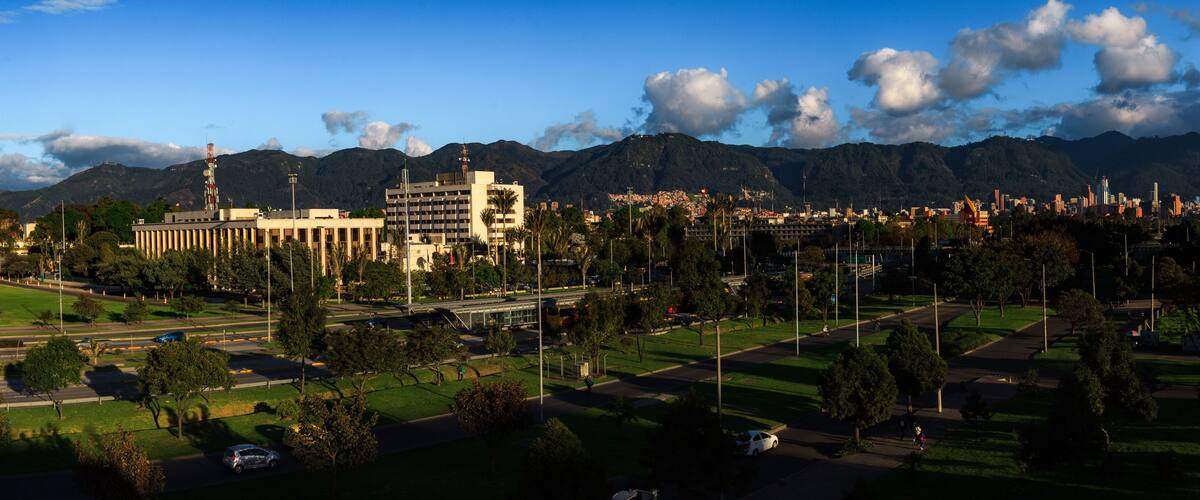 Un paisaje urbano con montañas al fondo, una mezcla de edificios modernos y antiguos, y una zona verde de parque.