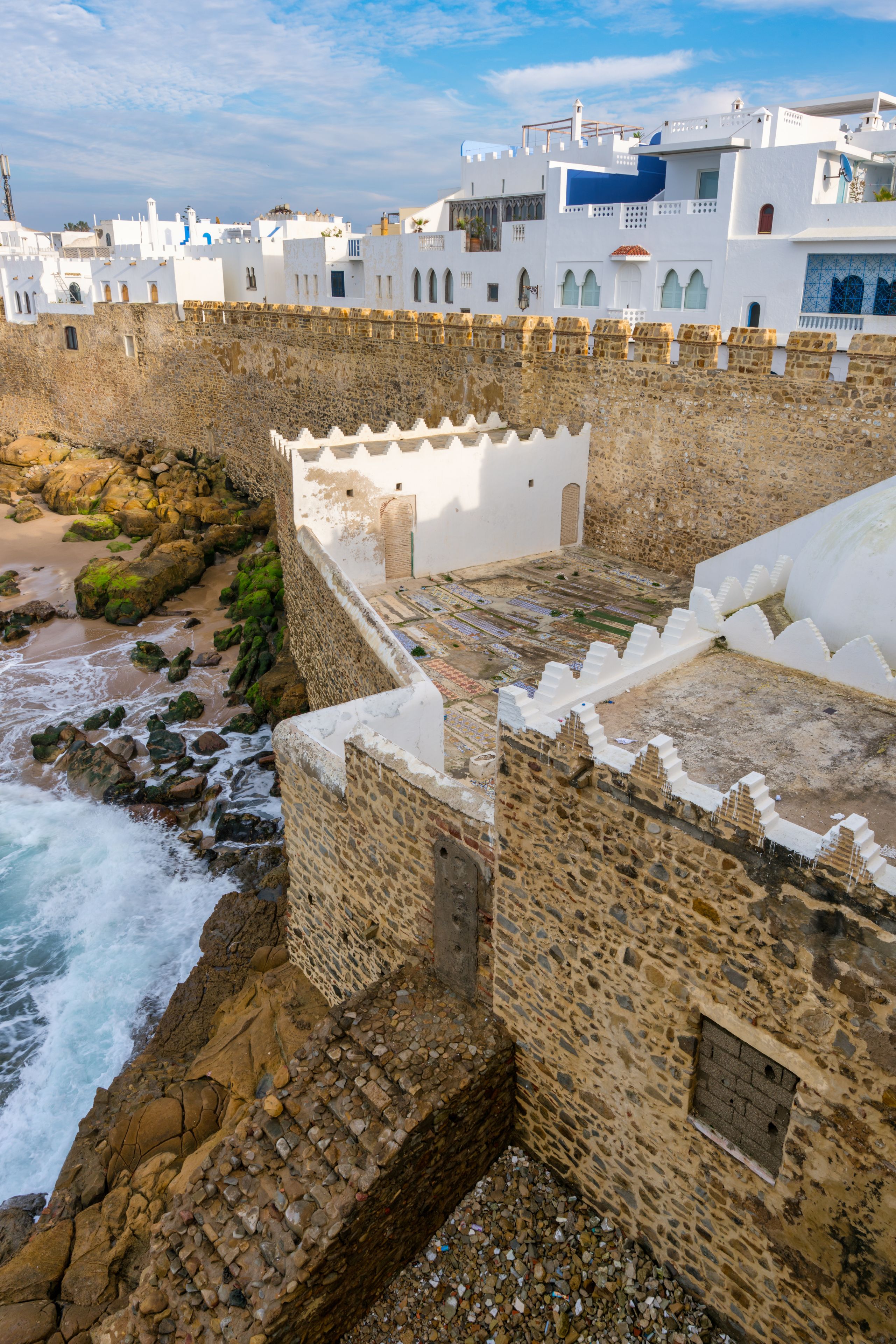 View of the historic stone ramparts and white-washed buildings of the Asilah medina overlooking the rocky Atlantic coastline in Asilah, Tangier-Tetouan-Al Ho, Morocco.