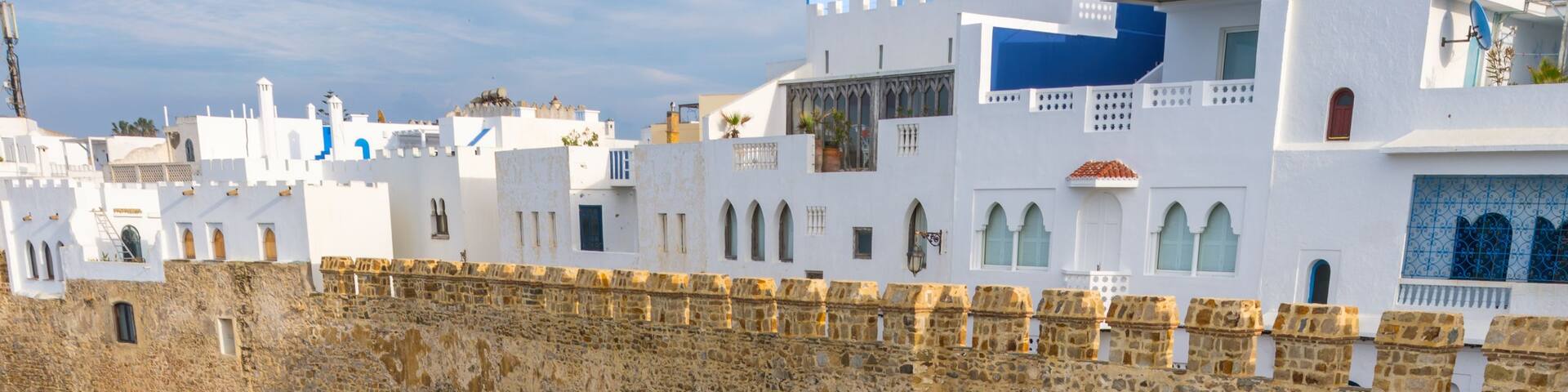 A coastal fortification with white walls and a stone base, overlooking the sea with waves crashing against the rocks.