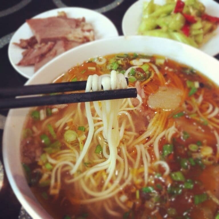 A bowl of noodle soup with chopsticks, garnished with green onions and possibly other vegetables.