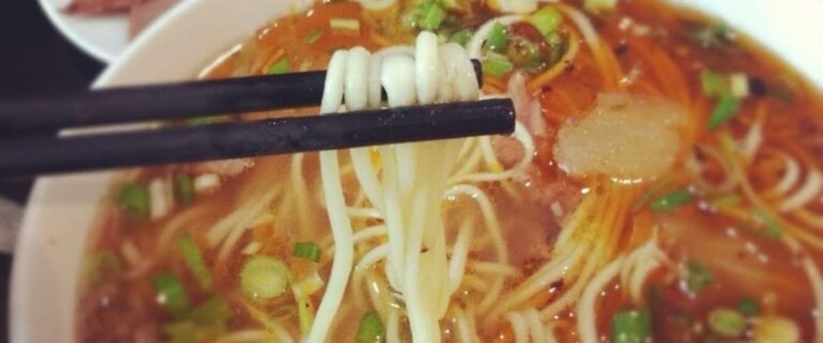 A bowl of noodle soup with chopsticks, garnished with green onions and possibly other vegetables.