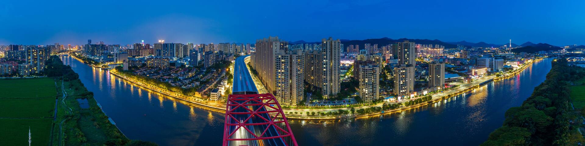 A cityscape at night with a river curving through it, illuminated buildings, and a prominent red bridge structure.