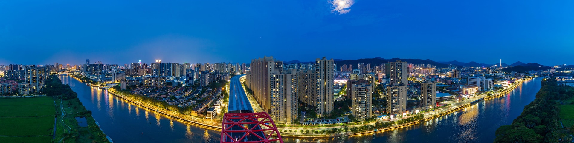 Uma paisagem urbana noturna com um rio serpenteando, edifícios iluminados e uma ponte vermelha imponente.