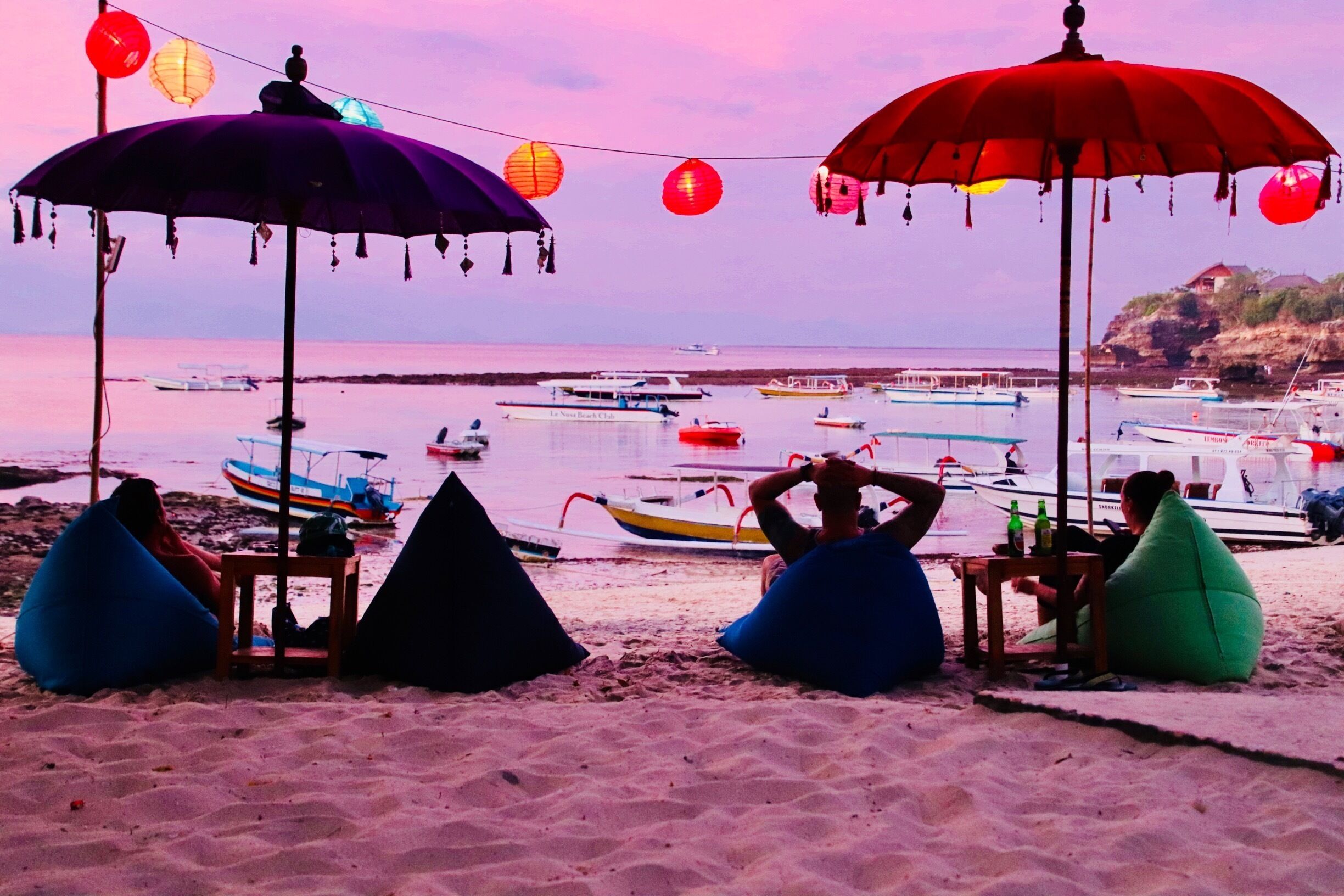Una playa con sombrillas de colores, pufs y barcos en el agua.