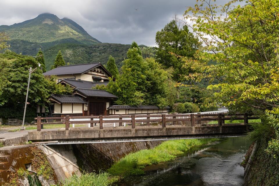 A traditional Japanese house by a river, surrounded by lush greenery and mountains.