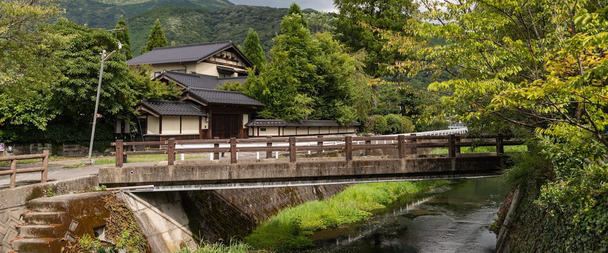 A traditional Japanese house by a river, surrounded by lush greenery and mountains.