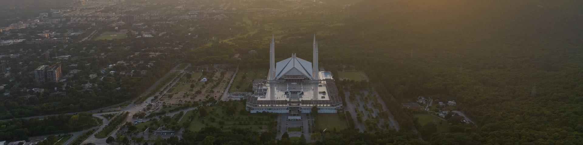 An aerial view of a large mosque with two minarets, surrounded by a cityscape and forested hills.