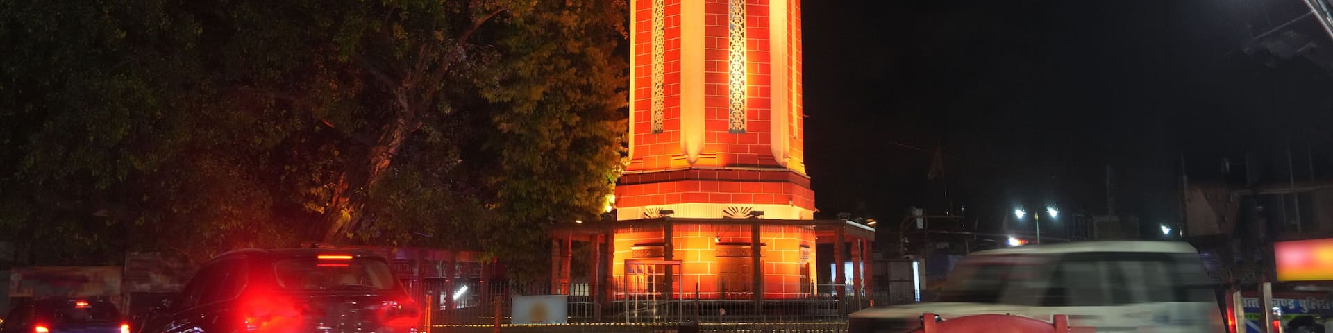 Night view of the illuminated Dehradun Clock Tower in Uttarakhand, India, glowing with warm architectural lighting against a dark sky. A landmark city scene featuring historic tower architecture